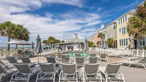 A poolside area with lounge chairs, umbrellas, and a building with balconies. - Stay @ The Grand in Wild Dunes (Isle Of Palms)