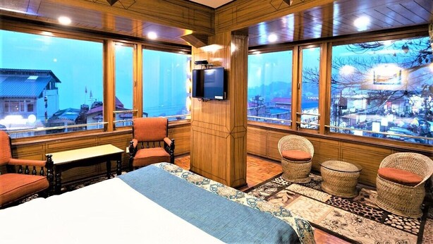 Une chambre d’hôtel avec un grand lit, deux fauteuils, une petite table et un téléviseur. La chambre est dotée de grandes fenêtres offrant une vue sur un paysage enneigé.