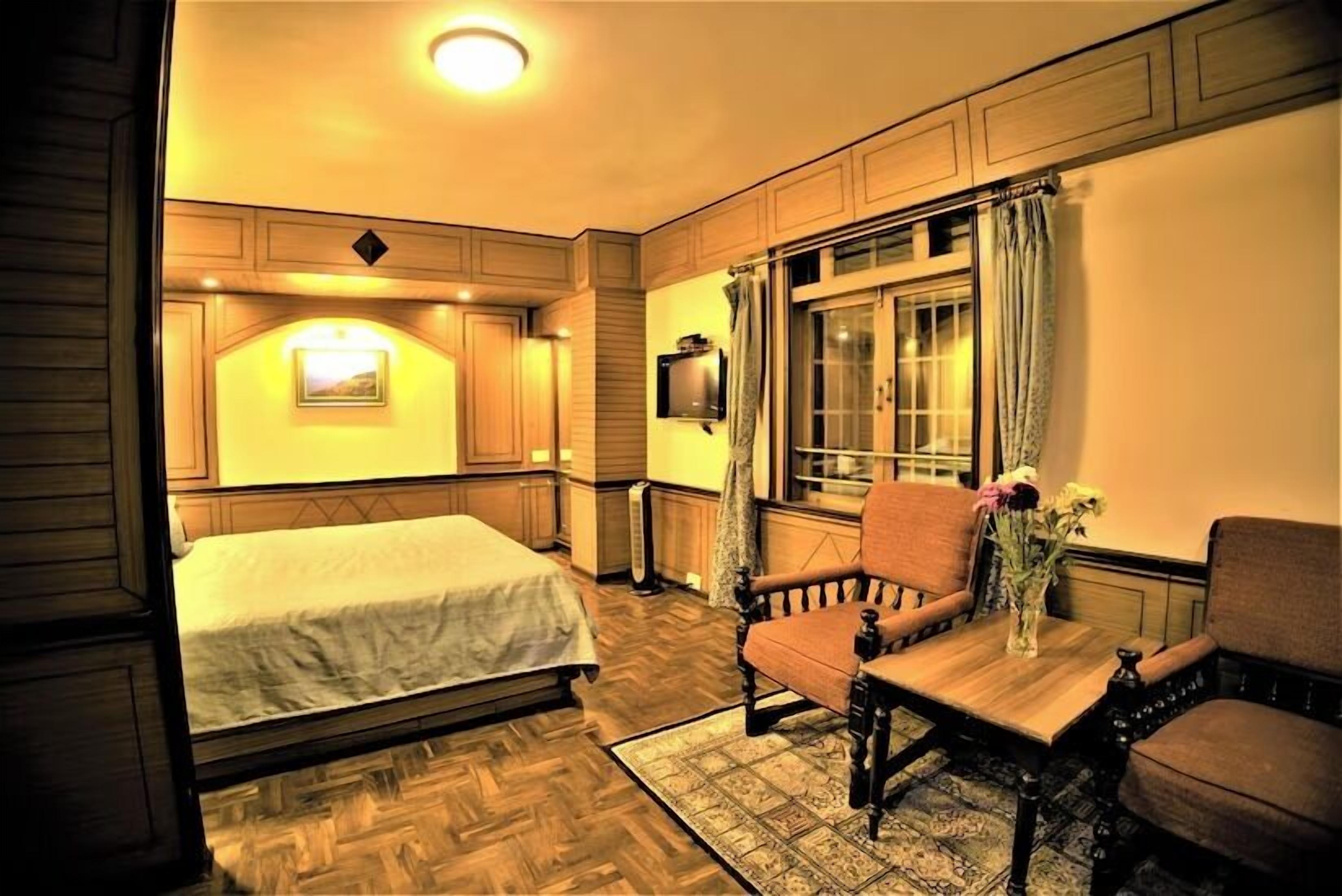 A hotel room with a bed, two armchairs, a small table with a vase, a window with curtains, and a mounted television.