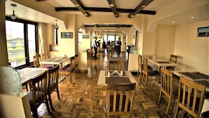 Un restaurant avec des planchers, des tables et des chaises en bois. On aperçoit des personnes au fond de la pièce, et la salle a un plafond haut avec des poutres apparentes.