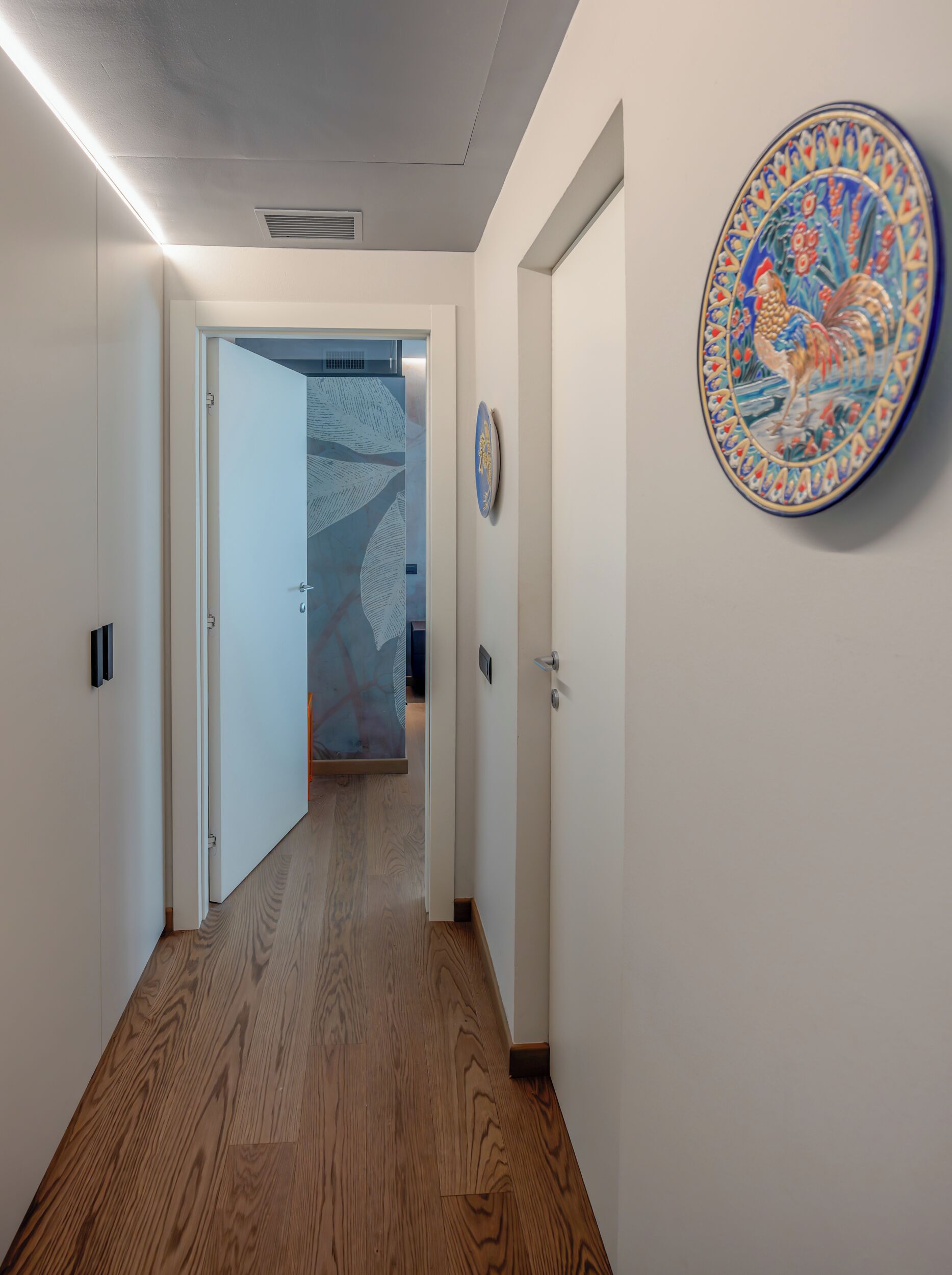 Un pasillo con suelo de madera, una puerta con placa decorativa y un papel tapiz estampado.