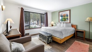 Hotellihuone, jossa on sänky, työpöytä, tuoli, pieni pöytä, lamppu ja ikkuna, jossa on verhot.