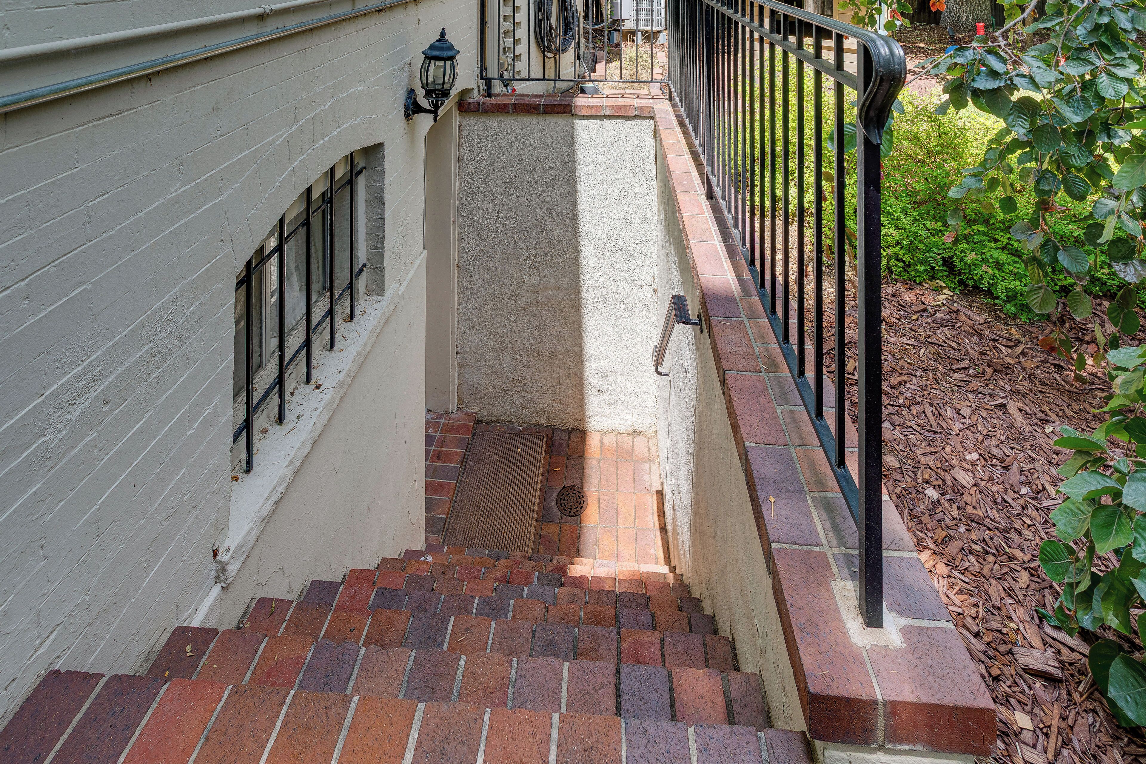 A narrow stairway with red brick steps leading to a white wall with a window and a black metal railing.
