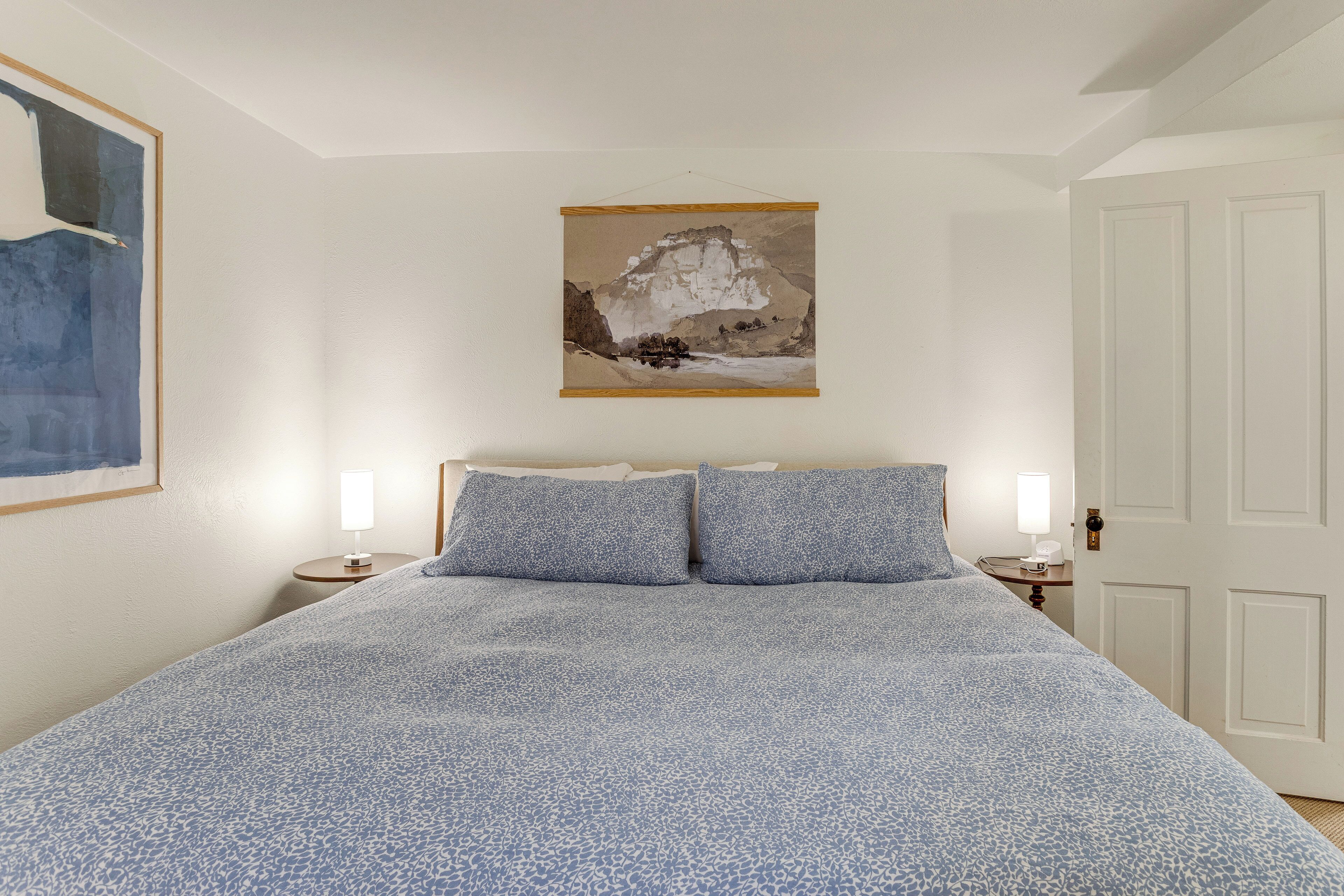 A bedroom with a bed, two bedside tables with lamps, a door, and a painting on the wall.