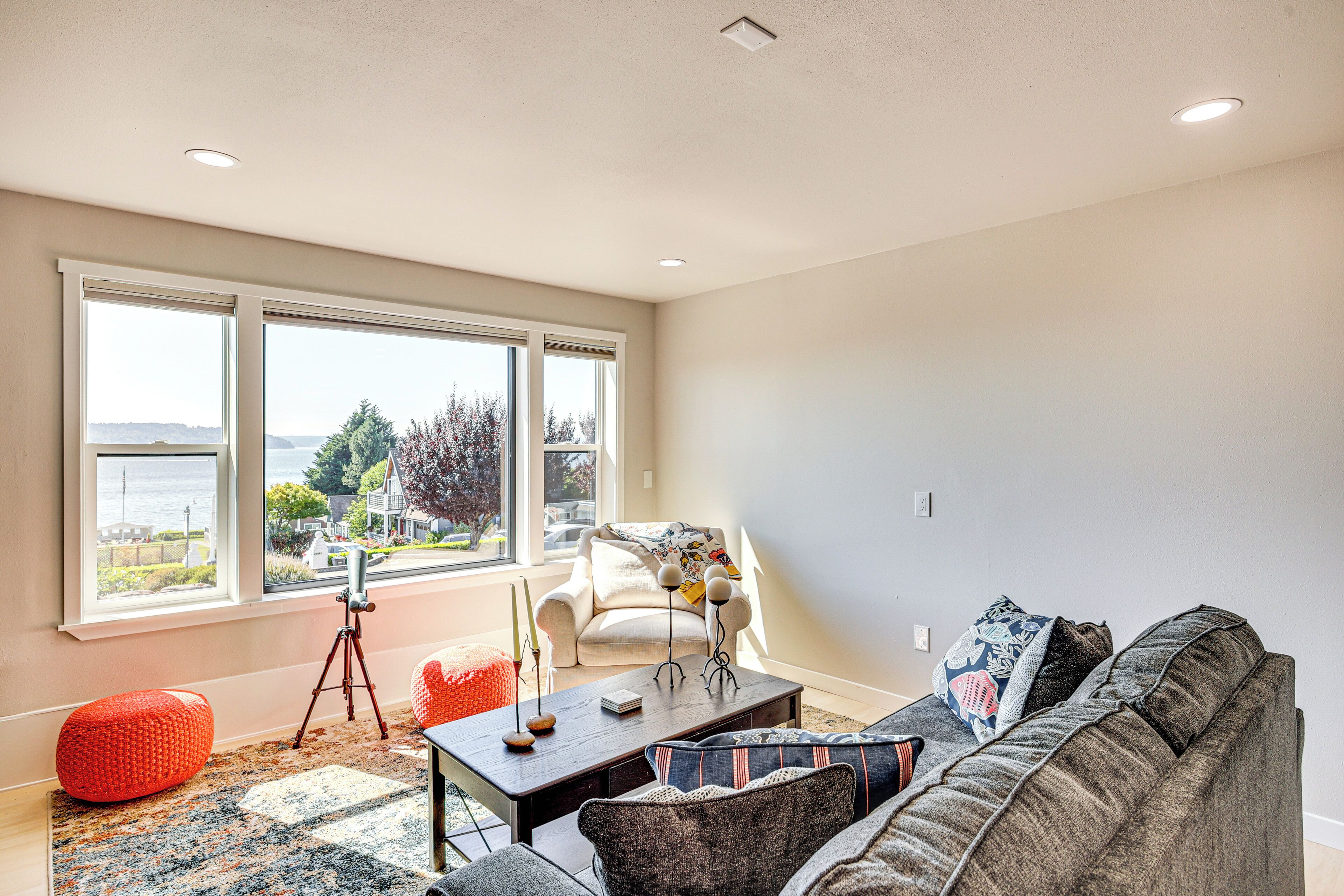 Un salon avec une grande fenêtre, un canapé, une table basse et un pouf rouge.