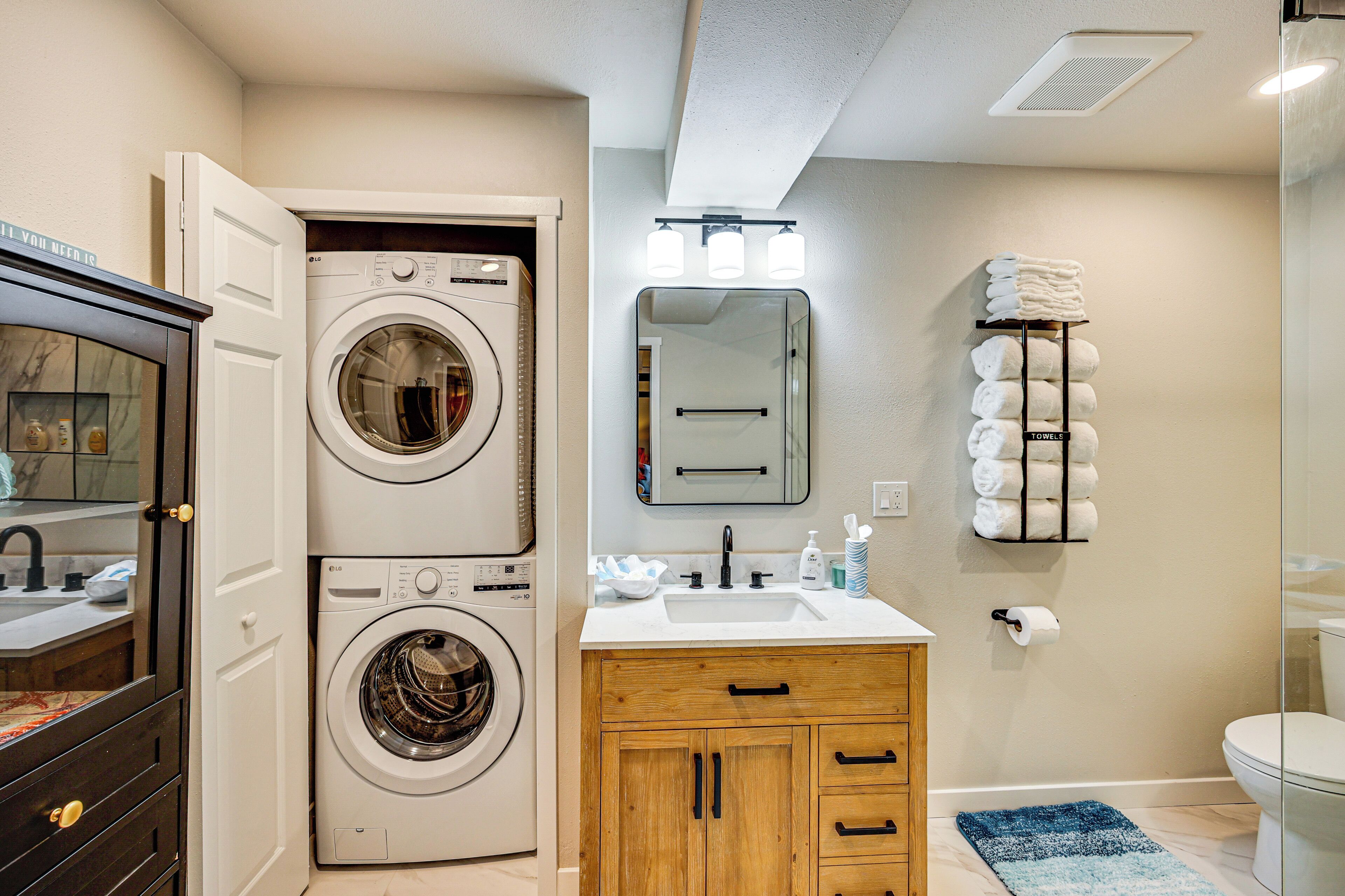 A laundry room with two stacked front-loading washing machines, a wooden cabinet with drawers, a sink with a mirror above it, and a towel rack with towels.