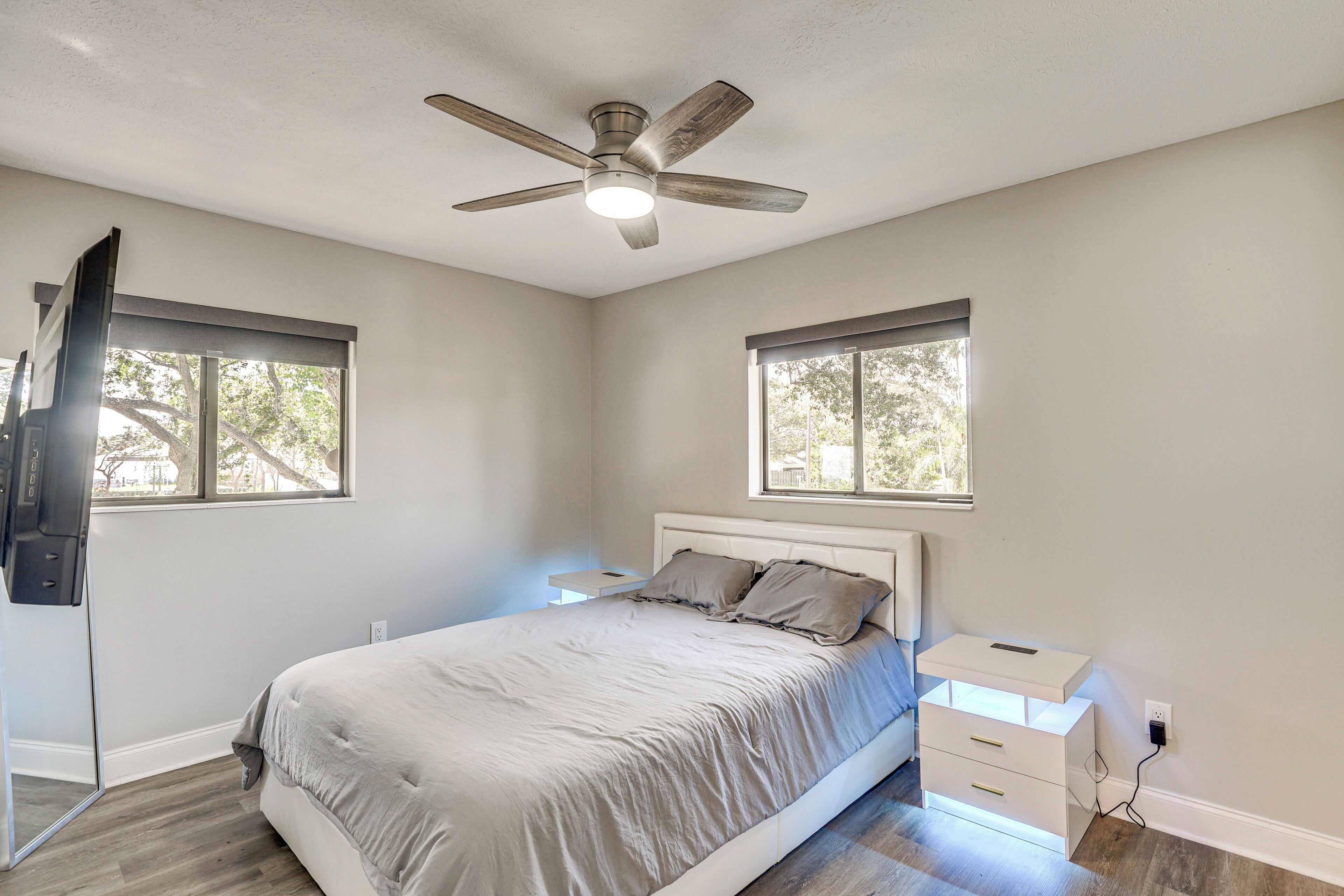 Une chambre à coucher avec un lit, une table de chevet, un ventilateur de plafond et deux fenêtres.