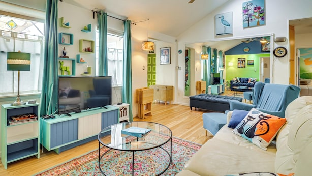 A living room with a TV, sofa, and coffee table. There are curtains, a ceiling fan, and artwork on the walls. - Outdoor Bar & Large Pool: Retro ABQ Retreat! (Albuquerque)