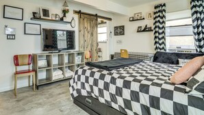 A bedroom with a checkered bedspread, a television, a wooden chair, a shelf with decorative items, and a window with curtains. - Outdoor Bar & Large Pool: Retro ABQ Retreat! (Albuquerque)