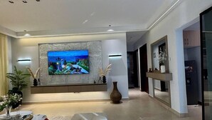A modern living room with a large flat-screen TV mounted on a textured wall, a beige sofa with red cushions, a round coffee table, and a tall vase with white flowers. - Ace Homes Bubga 3BR (Kampala)