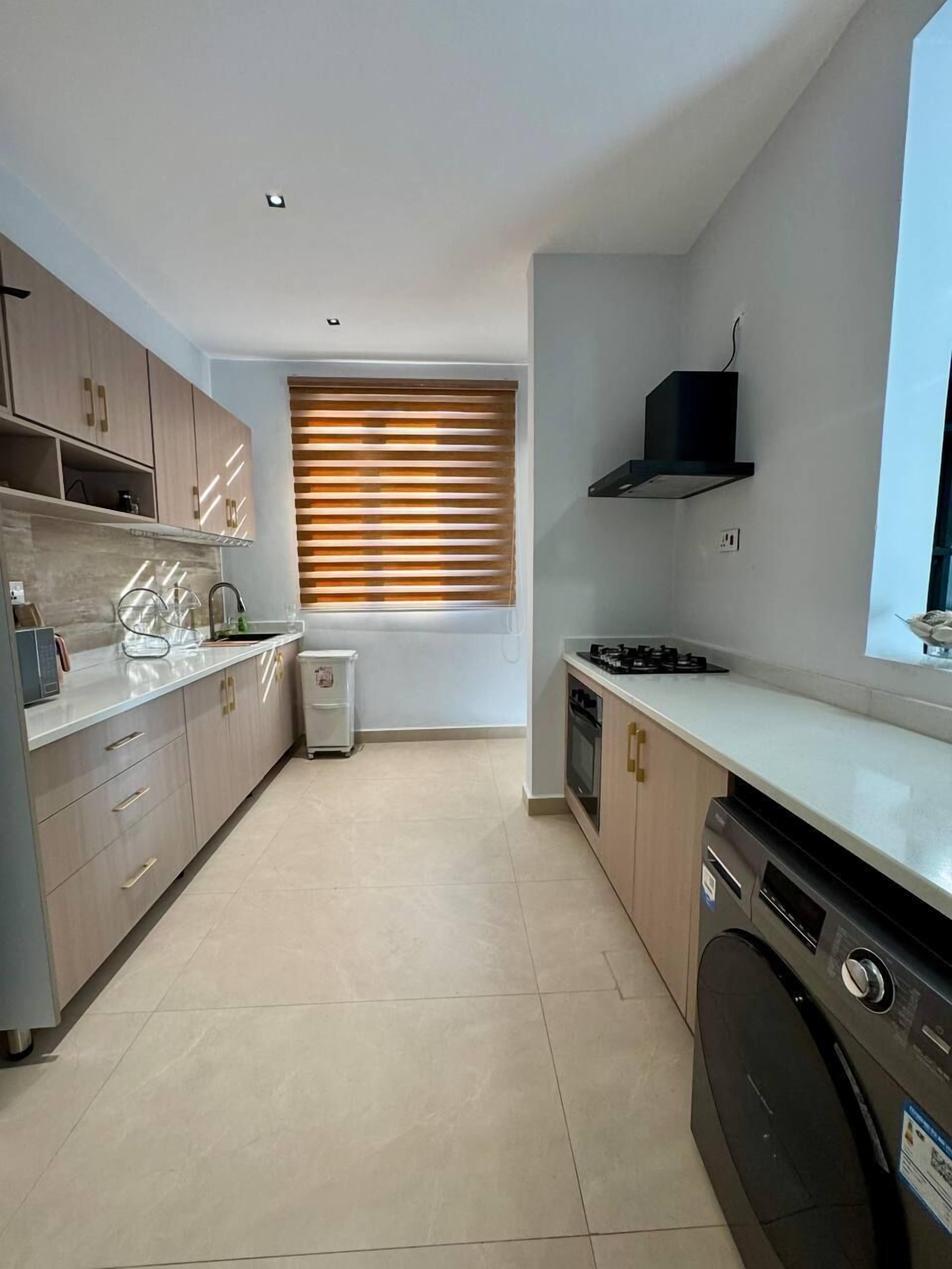 A modern kitchen with light wood cabinets, a built-in dishwasher, a stove, and a window with blinds.