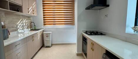 A modern kitchen with light wood cabinets, a built-in dishwasher, a stove, and a window with blinds.