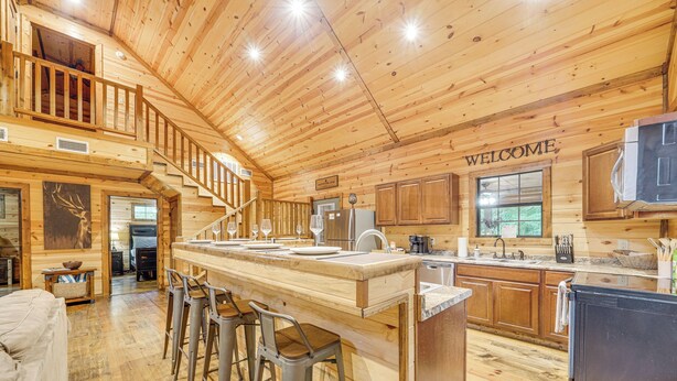 A spacious wooden cabin with a central bar area, a staircase, and a kitchen with wooden cabinets and a refrigerator. - Pets & Families Welcome: Secluded Broken Bow Cabin (Broken Bow)