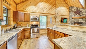 A spacious kitchen with wooden walls, granite countertops, stainless steel appliances, and a stone fireplace. - Pets & Families Welcome: Secluded Broken Bow Cabin (Broken Bow)