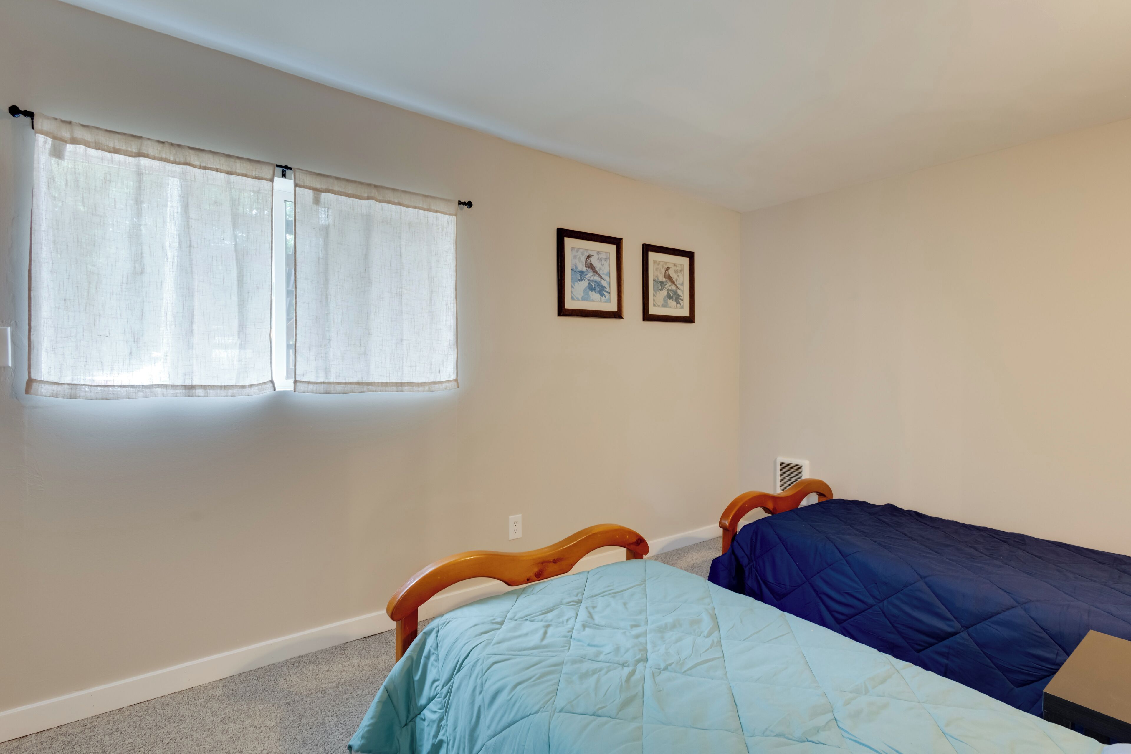 A bedroom with two single beds, a window with sheer curtains, and two framed pictures on the wall.
