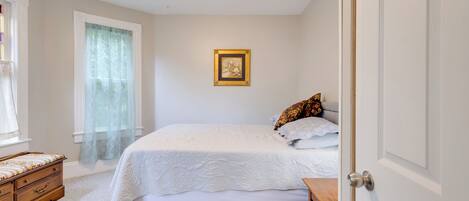 Un dormitorio con cama, un banco de madera, un pequeño tocador y una ventana con cortinas traslúcidas.