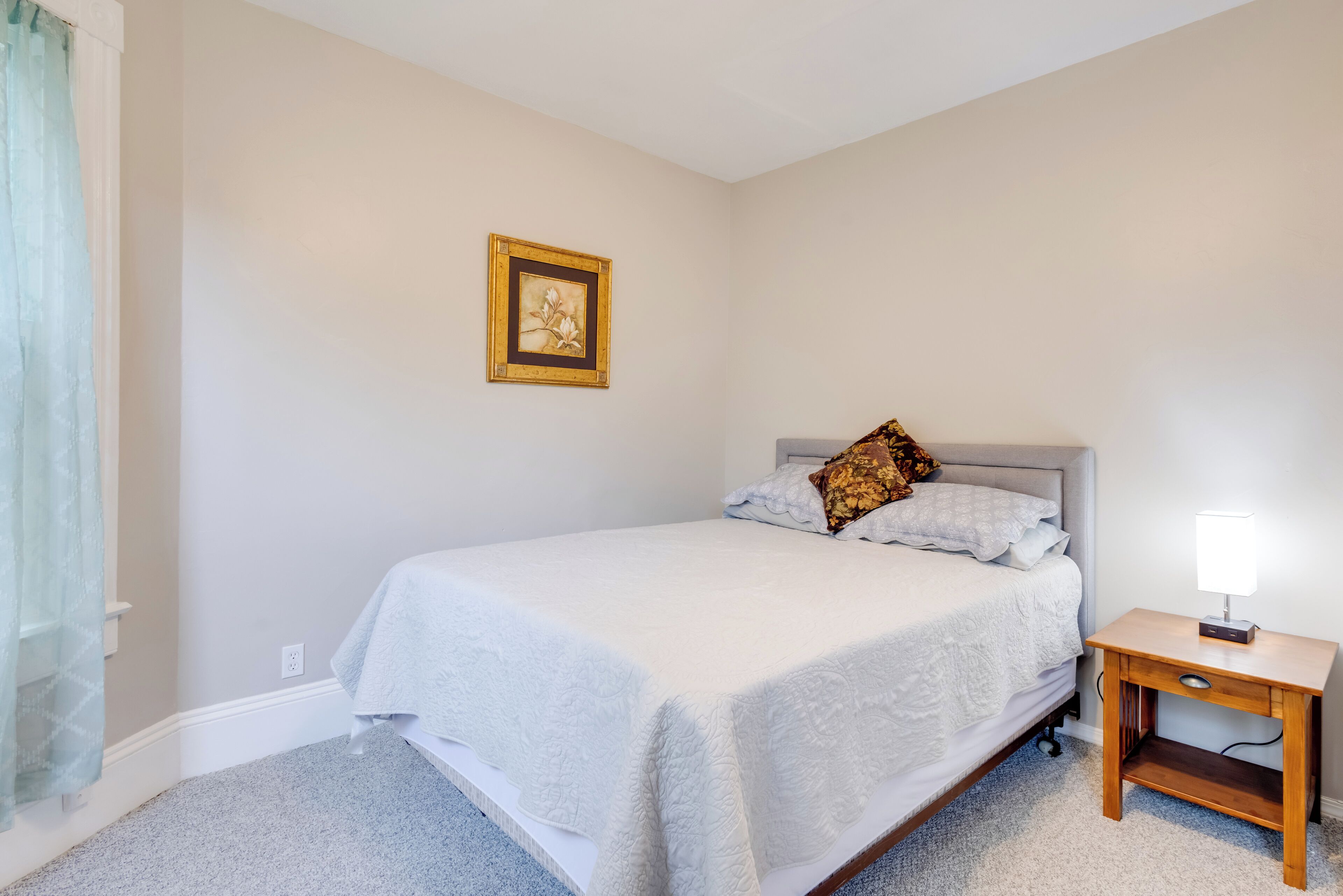 A bedroom with a bed, a bedside table with a lamp, a framed picture on the wall, and a window with a curtain.
