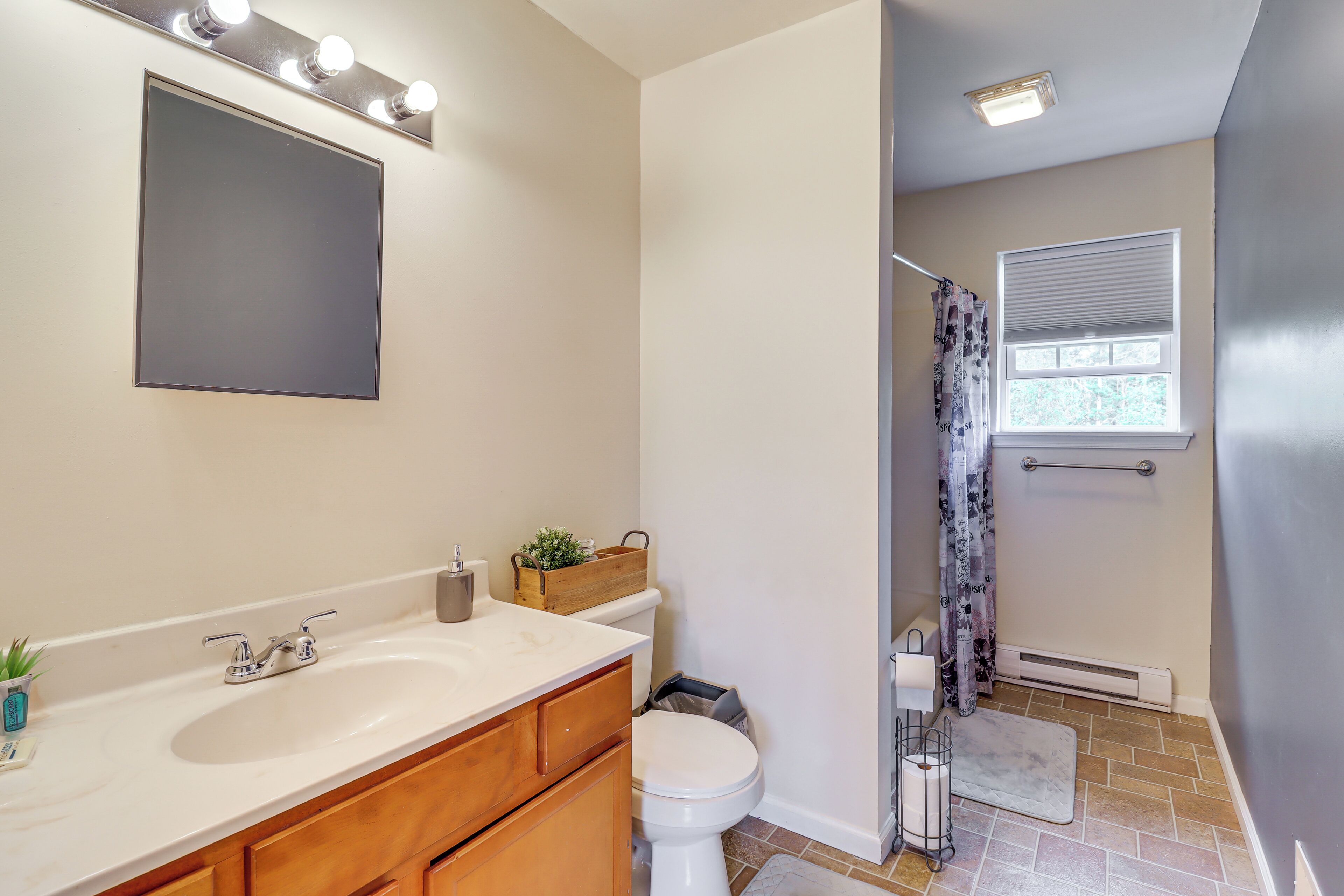 A bathroom with a white sink, wooden vanity, and a mirror with lights. There is a window with blinds and a shower area with a curtain.