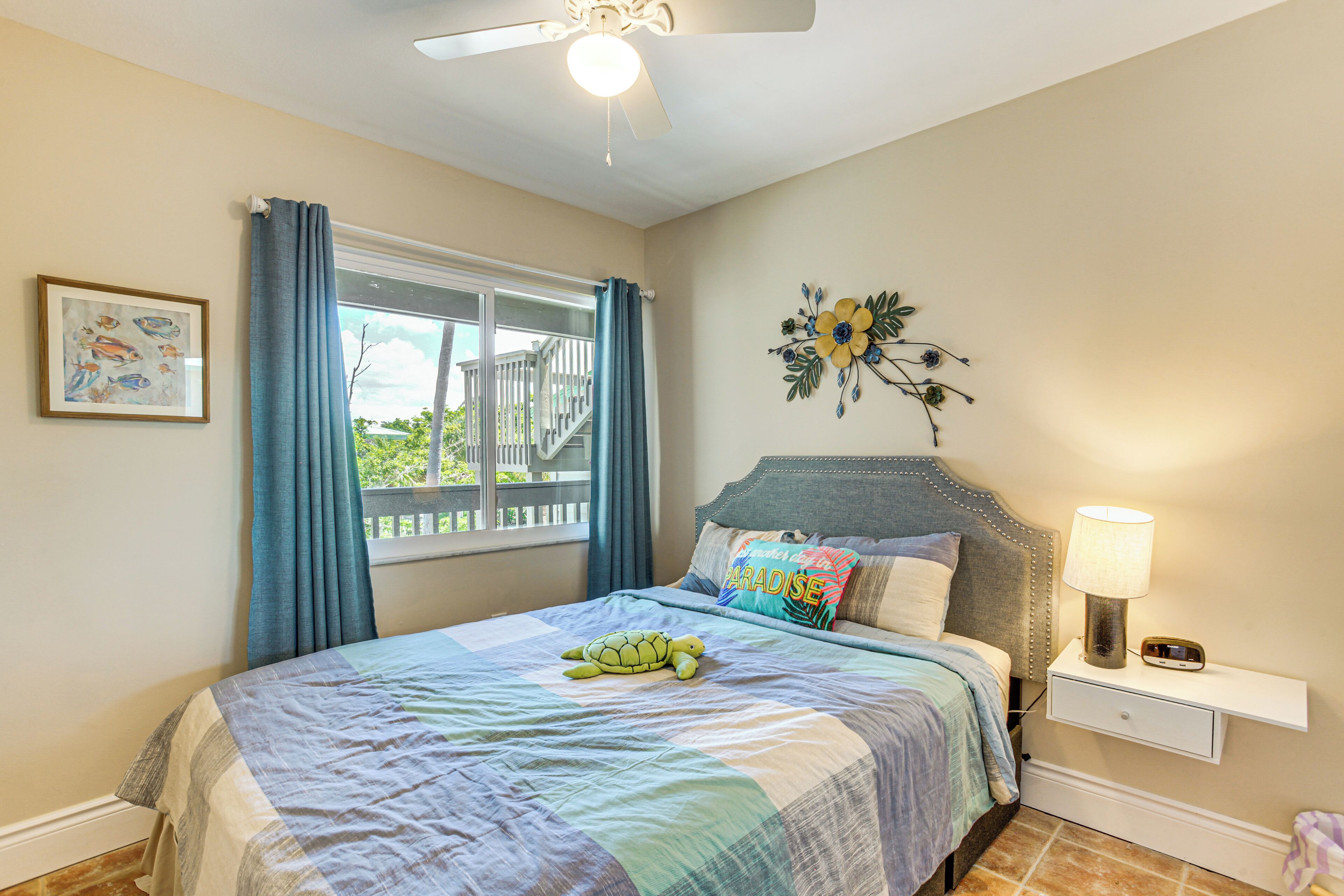 Une chambre à coucher comprenant un lit, une table de chevet, une lampe, un ventilateur de plafond, une fenêtre avec des rideaux et un élément de décoration murale.
