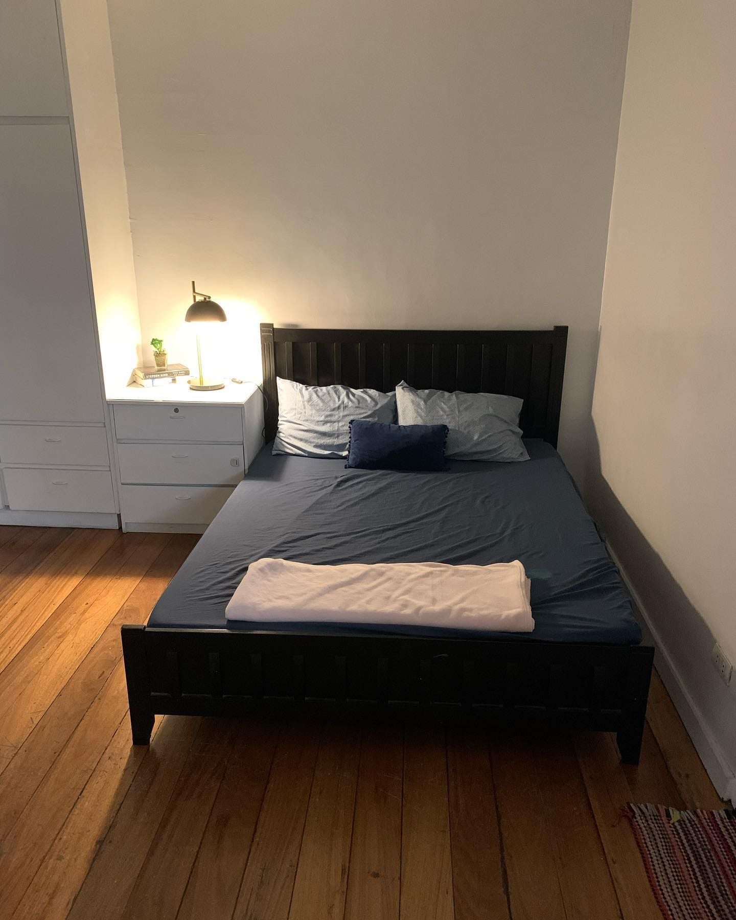 Een bed met een donker hoofdbord, een nachtkastje met een lamp en een kleine plant.