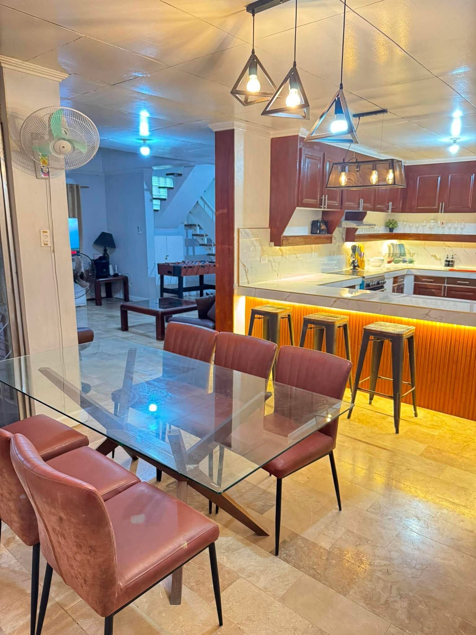 Une cuisine moderne avec une table à manger en verre et des chaises rouges, un coin bar avec des tabourets et des luminaires suspendus.