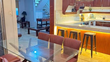 Une cuisine moderne avec une table à manger en verre et des chaises rouges, un coin bar avec des tabourets et des luminaires suspendus.