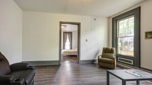 A room with a dark wooden floor, a black leather sofa, a wooden coffee table, a beige armchair, and a large window with a view of trees. - Walk to Dtwn Bath: Apt Near Keuka Lake & Wine Trl (Bath)