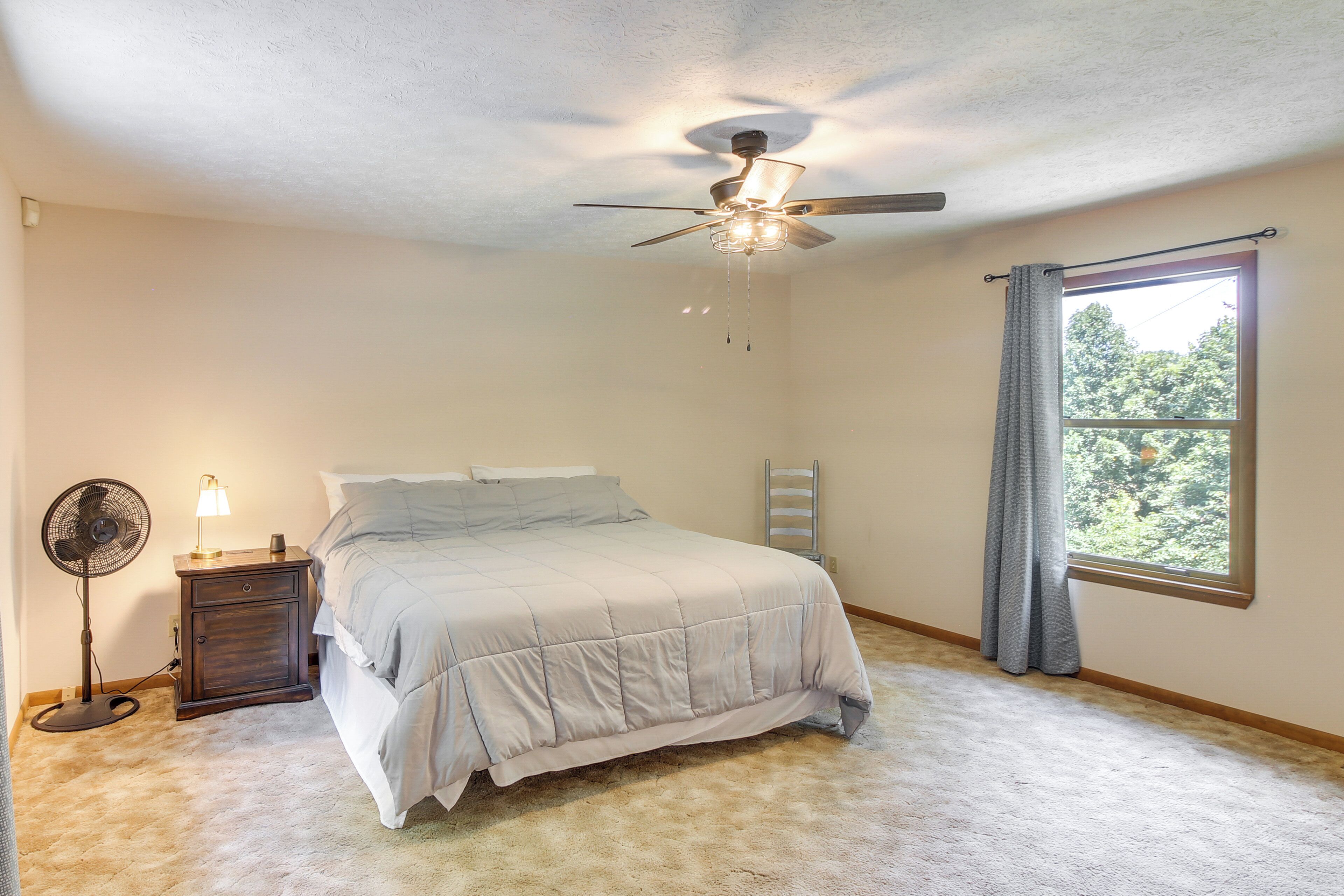 A bedroom with a bed, a ceiling fan, a nightstand with a lamp, a window with curtains, and a view of trees outside.