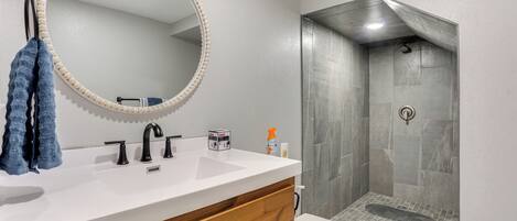 A bathroom with a white sink, wooden vanity, a round mirror, a towel, and a shower area with tiled walls.