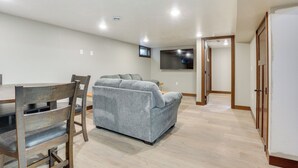 Una sala de estar moderna con un sofá gris, una mesa de comedor de madera y sillas. Hay un televisor de pantalla plana montado en la pared y una puerta que da a otra habitación.
