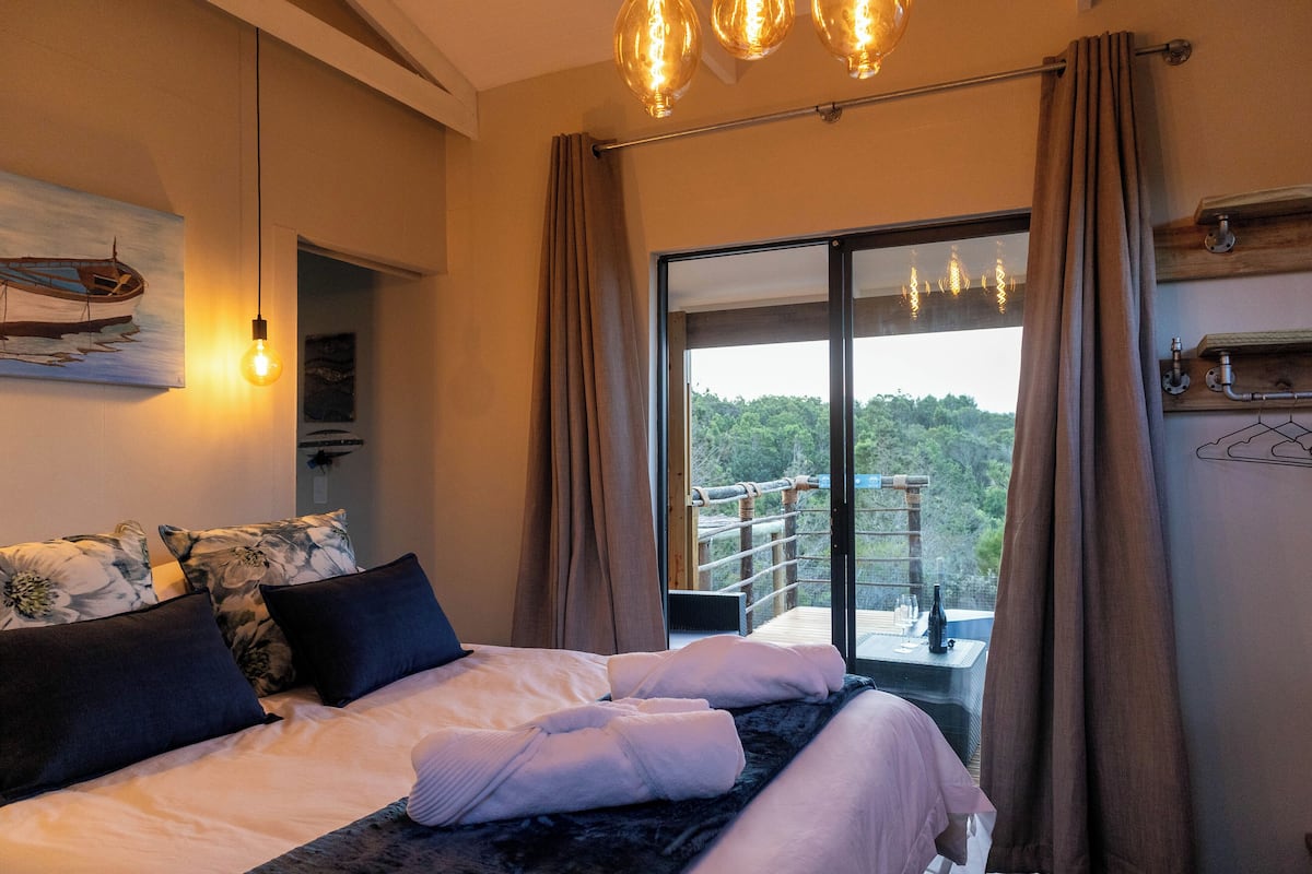 A bedroom with a large bed, a painting of a boat, a hanging light fixture, and a view of a balcony with a railing.