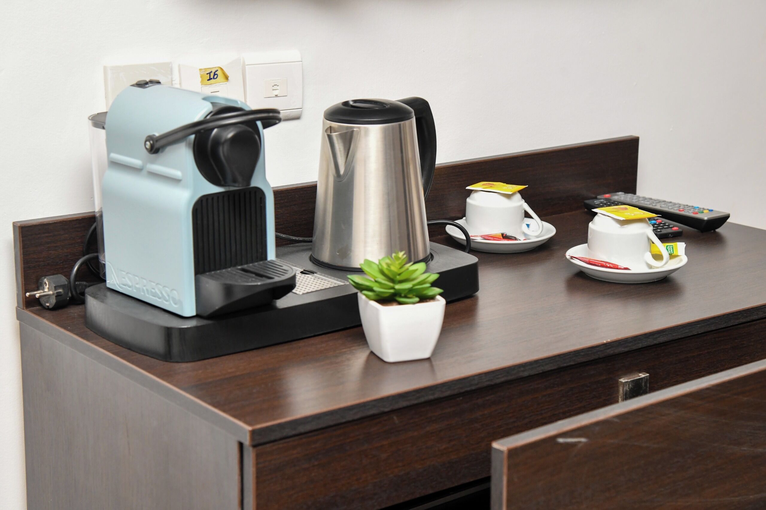 Uma cozinha compacta com cafeteira, chaleira, uma pequena planta em vaso e controle remoto sobre um balcão de madeira.