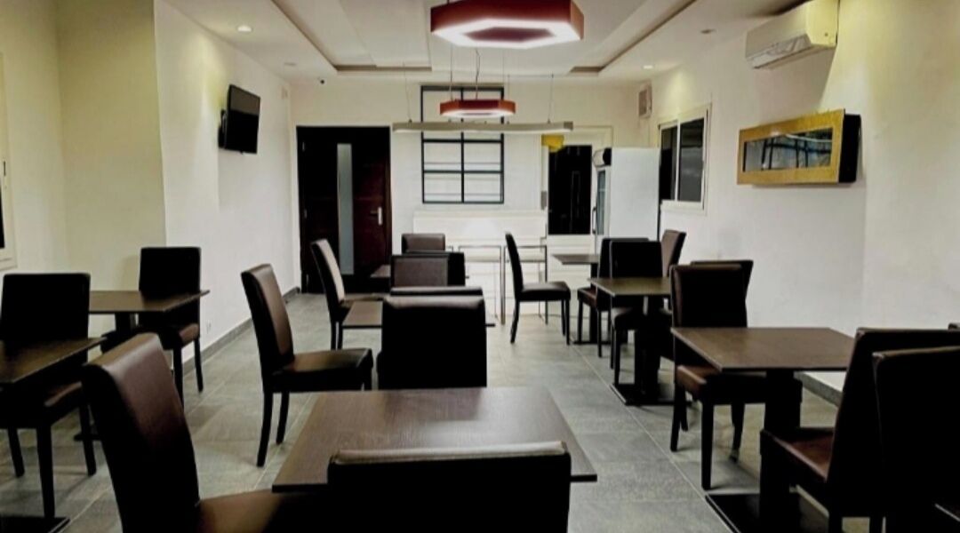 A spacious dining area with multiple tables and chairs, a flat-screen TV mounted on the wall, and a large window providing natural light.