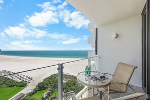 A balcony with a view of a beach, a round table with two bottles, and a chair. - 18th Floor Beachfront Condo | Pool, Gym & Grills (Marco Island)