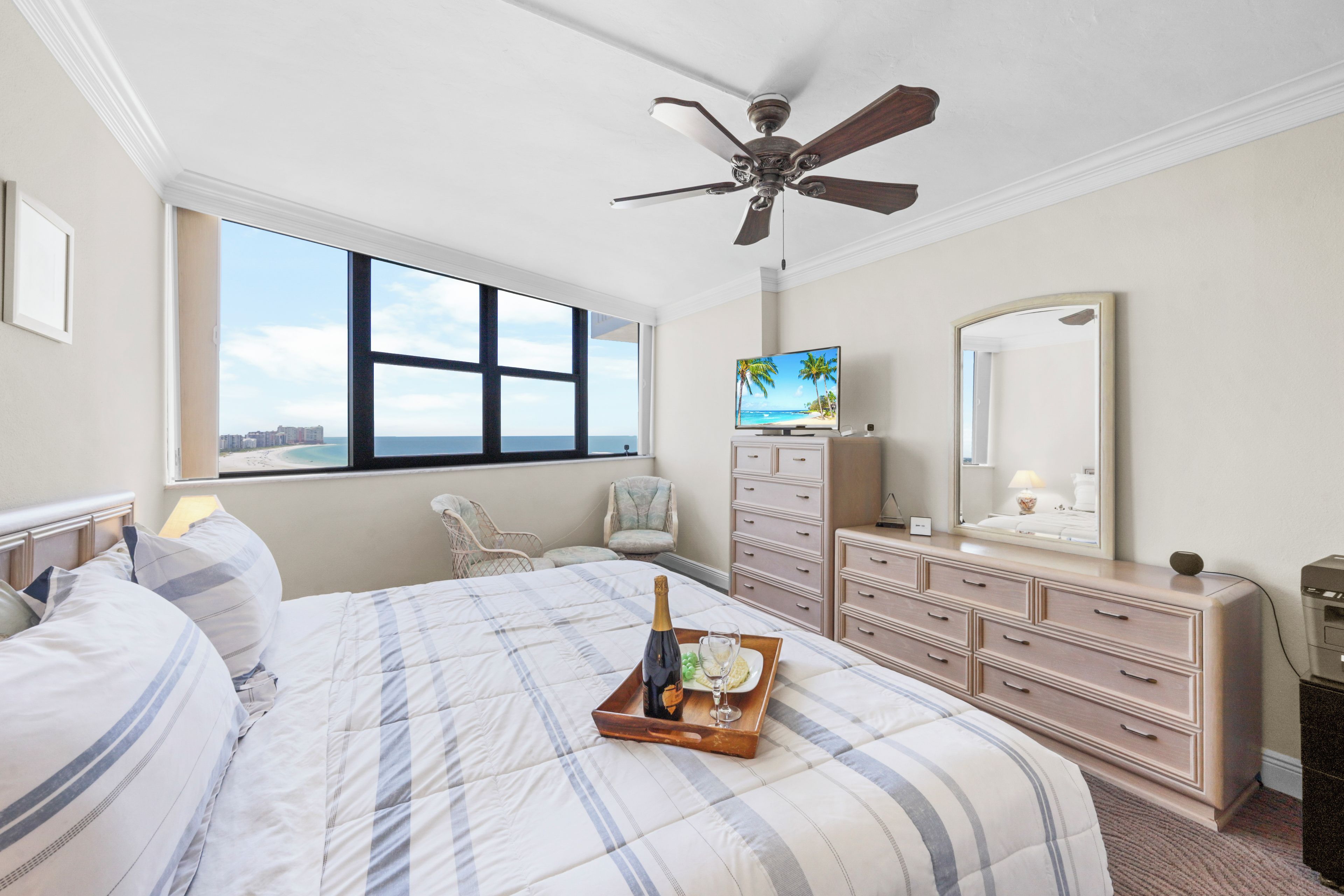 Een hotelkamer met een groot bed, een plafondventilator, een televisie, een dressoir en een raam met uitzicht op de oceaan.