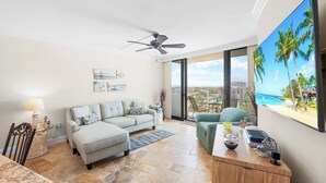 Amplia sala de estar con un gran ventanal con vista a la playa, un ventilador de techo, un sofá, una mesa de comedor y un escritorio con una computadora.