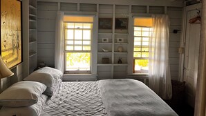 Un dormitorio con cama, dos ventanas con cortinas, una estantería con objetos decorativos y un cuadro en la pared.