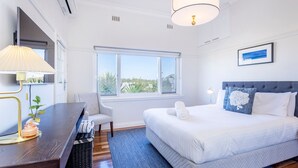 A modern hotel room with a large bed, a desk with a lamp, a chair, and a window offering a view of greenery.