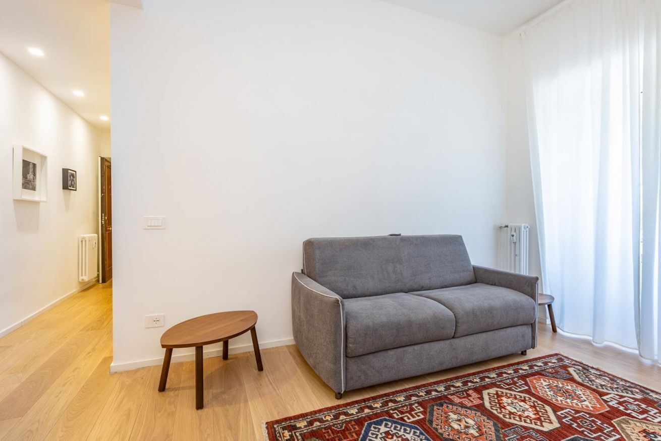 A living room with a grey sofa, a small wooden table, a patterned rug, and a white wall with a door and radiator.