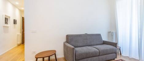 A living room with a grey sofa, a small wooden table, a patterned rug, and a white wall with a door and radiator.