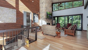 A spacious living area with a wooden staircase, a stone fireplace, and large windows offering a view of the outdoors.