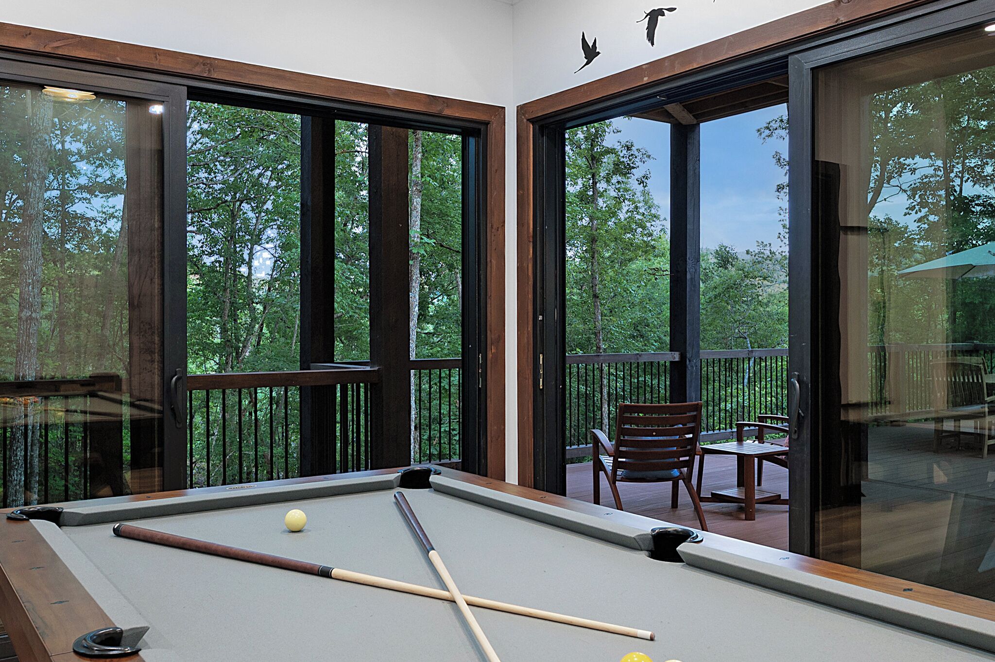 A pool table with a wooden frame and a pool cue, surrounded by a glass partition with a view of a wooden deck and outdoor furniture.