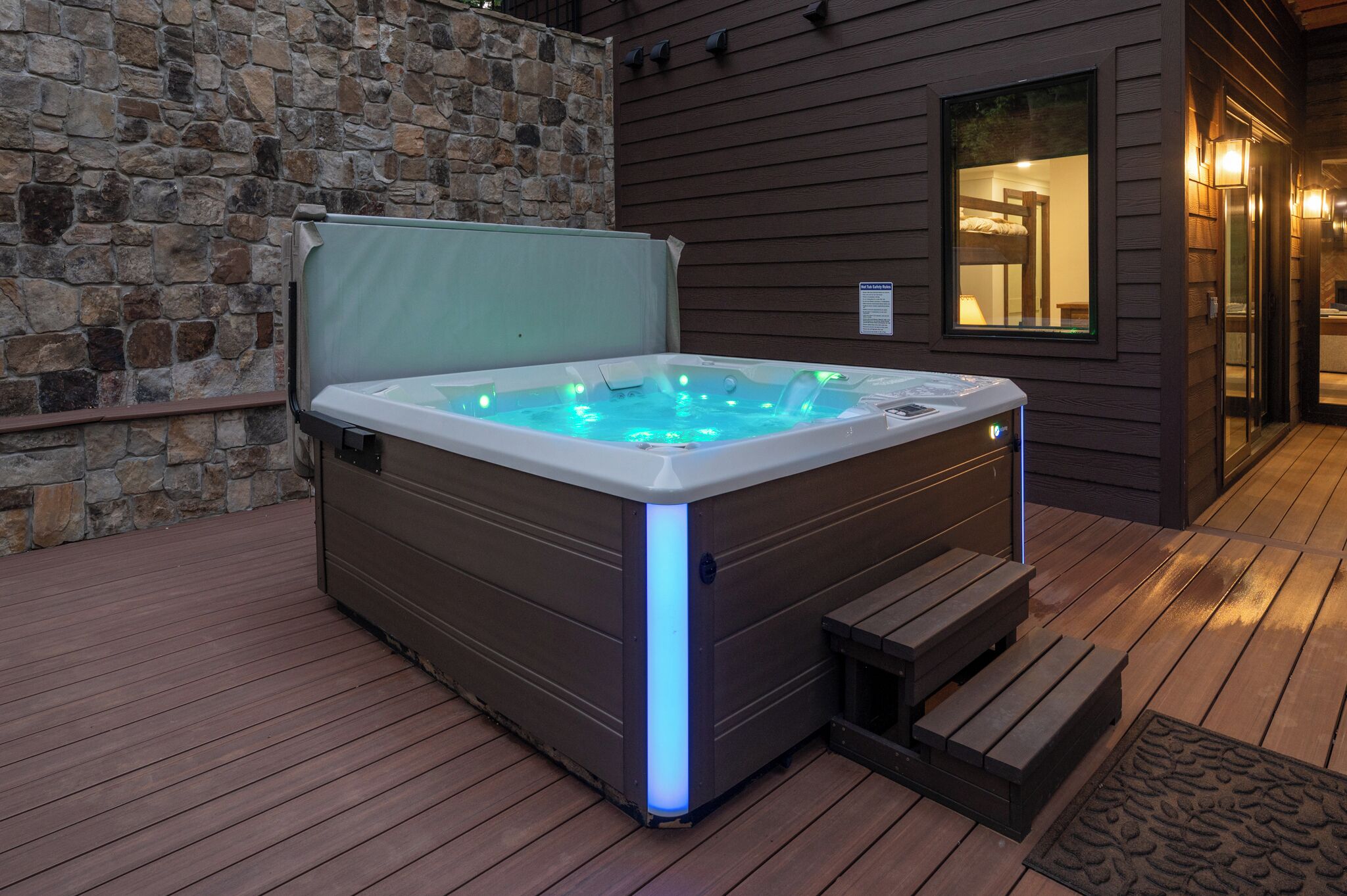 Un bain à remous doté d’une enceinte en verre, d’une terrasse en bois, d’un mur en pierre et d’une construction en bois avec une fenêtre.