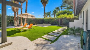 A well-maintained backyard with artificial grass, a stone pathway, and two lounge chairs under a striped umbrella.