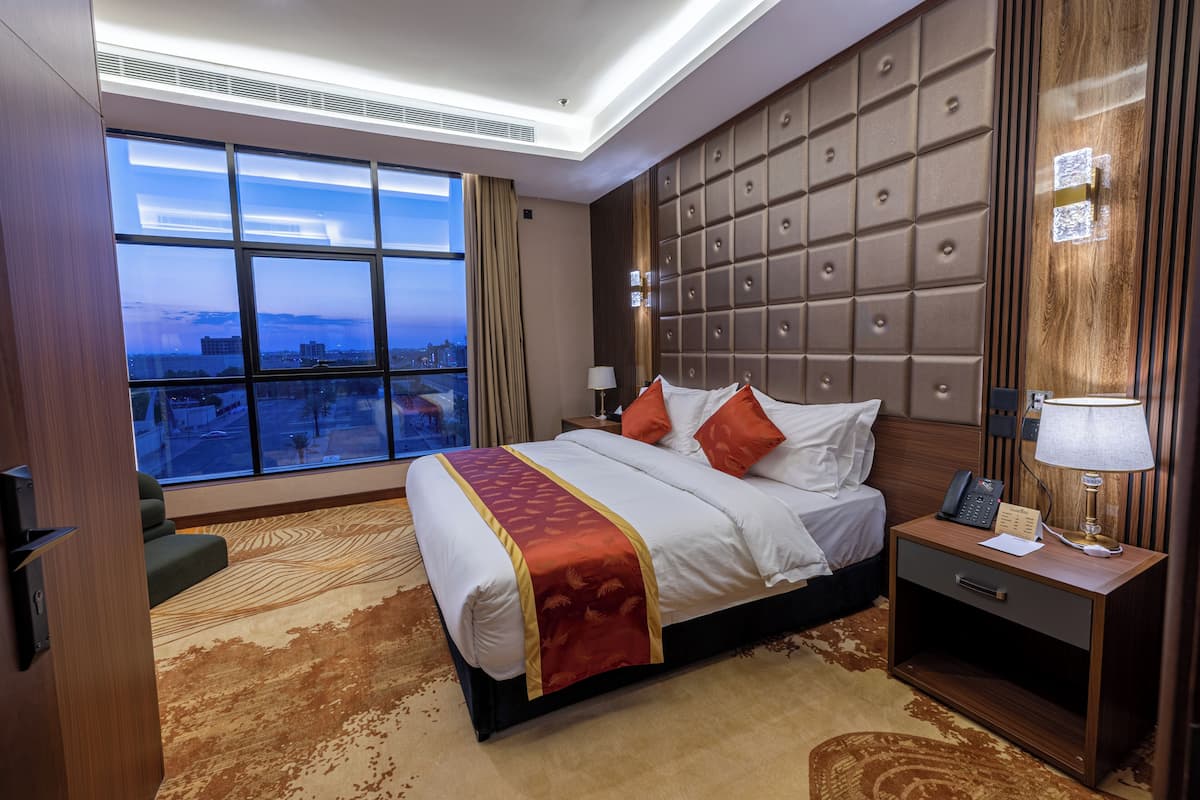 A hotel room with a large bed, a nightstand, a lamp, and a view of the city through large windows.