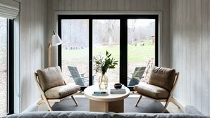 A room with a high ceiling, wooden walls, two leather chairs, a round table, a floor lamp, and a view of a grassy area through large windows.