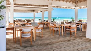 An outdoor dining area with wooden tables and chairs, overlooking a beach with clear blue water and a sailboat in the distance. - 2BR Beachfront Villa | Direct Access | Antigua Views (Urlings)