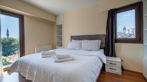 A neatly made bed with white linens and towels, a bedside table, and a view of a building through a window.