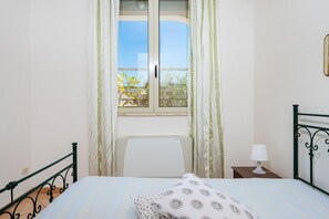 Ein Schlafzimmer mit Bett, einem Fenster mit Vorhängen, einem Nachttisch mit Lampe und Blick auf Bäume.