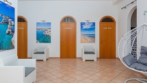 A hotel lobby with four wooden doors labeled 'Aguas Calientes,' 'Aguas Calientes,' 'Aguas Calientes,' and 'Aguas Calientes.' There are white wicker chairs and a white hanging chair with cushions.